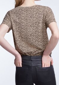 Tričko s krátkým rukávem v leopardím vzoru v béžové a hnědé barvě, s volným střihem. Modelka má na sobě černé kalhoty s zadními kapsami, ruce má opřené v bocích.