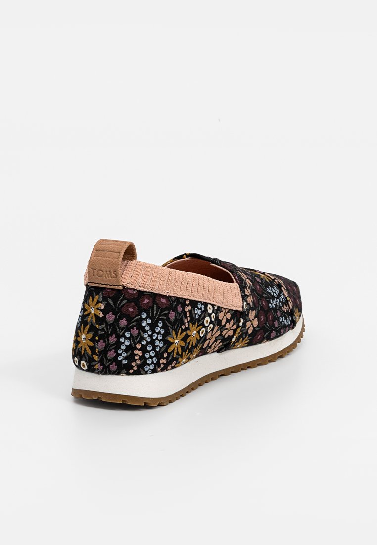 Zapato sin cordones con patrón floral, fabricado en tela negra, cinta elástica rosa y entresuela blanca. Cuenta con una suela de goma para mayor tracción.