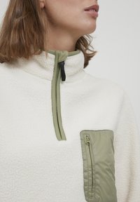 Fleece pulóver v krémovej farbe, s zeleným zipsom na golieri a zelenou zipsovou náprsní kapsou. Mäkká štruktúra s kontrastnou tkaninou.