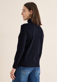 Granatowy, prążkowany top z długim rękawem z wysokim kołnierzem i bocznymi rozcięciami, noszony z niebieskimi dżinsami. Posiada dopasowany krój i gładką teksturę.