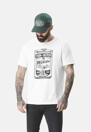 T-shirt blanc avec un imprimé graphique représentant un design de bidon d'huile vintage. Le modèle porte une casquette en maille verte, affichant le logo Von Dutch.