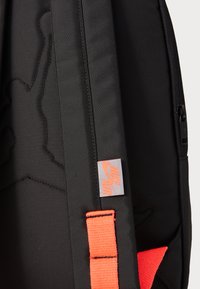 Sac à dos noir fabriqué en tissu texturé, avec une sangle orange et une étiquette avec le logo Nike Air. Fermeture éclair sur le côté et design en relief distinct visible.