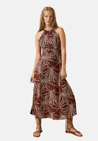 Vestido marrón sin mangas con patrón de hojas blancas, tejido ligero, hasta los tobillos y hombros al aire. Combinado con sandalias marrones de tobillo.