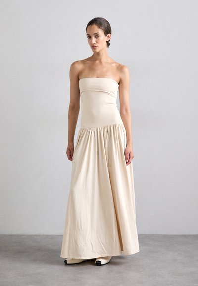 Robe maxi beige sans bretelles avec un corsage ajusté et une jupe froncée. Fabriquée en tissu doux, elle présente une texture lisse et une silhouette fluide.