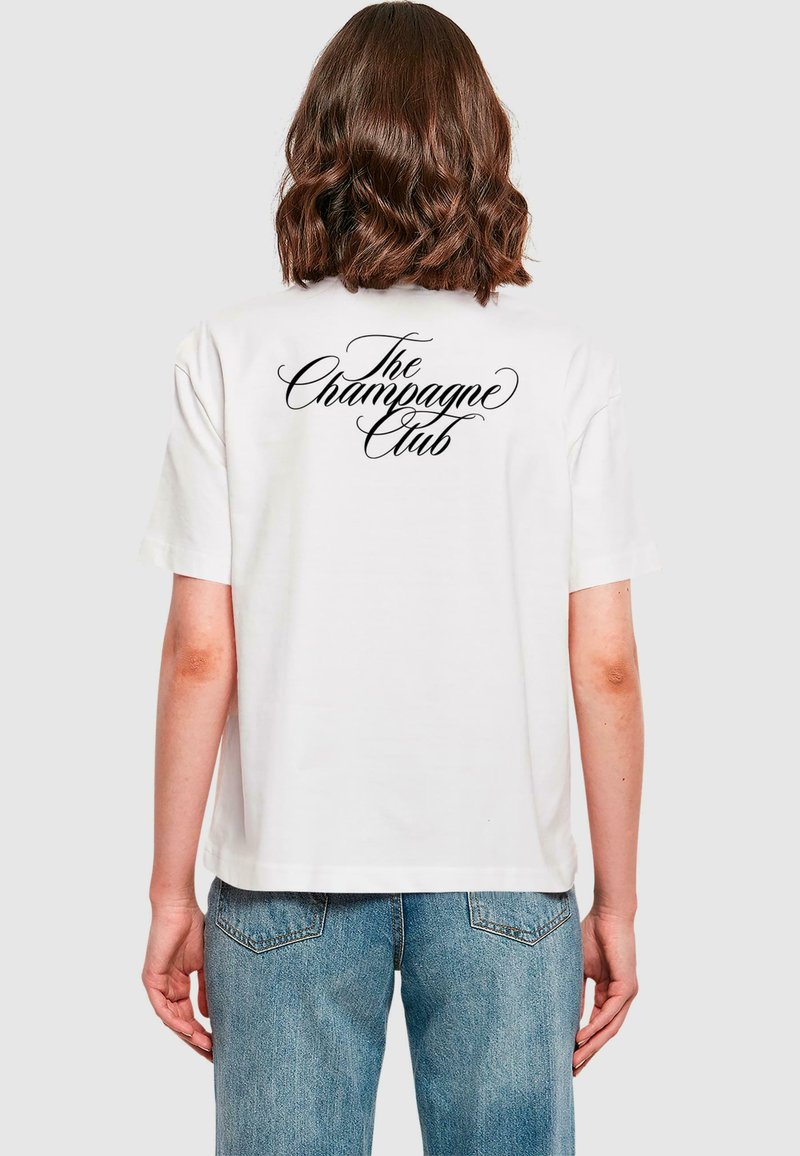 Vit t-shirt med en svart kursiv grafik som säger "The Champagne Club" på baksidan. Korta ärmar och avslappnad passform.