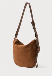 Brūna zamšādas plecu soma ar regulējamu jostu un zelta rāvējslēdzēja detalizāciju, pakārta pret baltu fona.