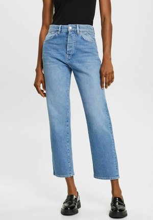 Kobieta ubrana w jasnoniebieskie, proste dżinsy z wysokim stanem, zestawione z czarnymi mokasynami i czarnym bezrękawnikiem, stojąca na jednolitym tle.