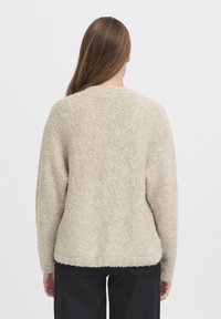Woman with long brown hair wearing a beige textured sweater and black pants, viewed from the back against a plain light background.