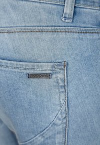 Los jeans de mezclilla de color azul claro cuentan con un bolsillo trasero con una pequeña etiqueta negra en relieve inscrita con "noppies". La tela presenta una textura de sarga clásica.