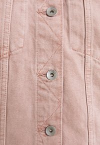 Chaqueta de denim de color rosa claro con botones a presión plateados, detalles de costura diagonal y un acabado de tela texturizada. Vista en primer plano del material.