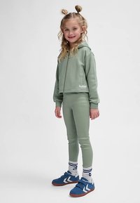 Groene geribbelde cropped hoodie met bijpassende legging en blauwe sneakers. Voorzien van ritssluiting, een logo en gelaagde sokken met strepen.