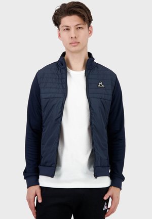 Chaqueta azul marino con parte superior acolchada, puños acanalados y cierre de cremallera en el frente. Logo en el lado izquierdo del pecho. Tejido suave con un diseño deportivo.