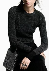 Schwarzes, schimmerndes Langarmoberteil mit geripptem Muster. Kombiniert mit einer schwarzen Tasche, die kreisförmige Verzierungen aufweist, und metallischen Ringen an den Fingern.