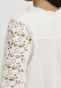 Blouse blanche en tissu texturé, col court à volants, et manches en dentelle blanche complexe avec motifs floraux et bords festonnés.