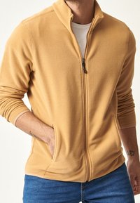 Giacca in pile con zip di colore giallo senape, caratterizzata da un colletto alto e tasche laterali. Il tessuto è morbido e strutturato, ideale per essere indossato a strati.