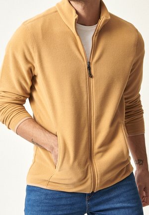 Chaqueta de forro polar con cremallera en mostaza, con un cuello alto y bolsillos laterales. La tela es suave y texturizada, ideal para llevar en capas.