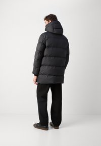 Schwarze Daunenjacke mit Kapuze, ausgestattet mit einem gesteppten Design, langen Ärmeln und einem leicht abgerundeten Saum. Hosen und Schuhe in dunklen Farben.