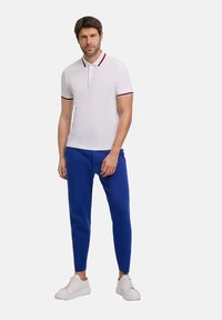 Biała koszulka polo z czerwono-granatowym paskiem przy kołnierzyku, w połączeniu z niebieskimi, dopasowanymi spodniami i białymi trampkami. Gładki materiał, wysmuklający krój.