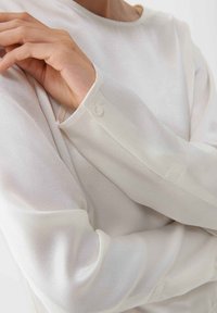Maglietta a maniche lunghe di un beige chiaro, caratterizzata da un tessuto liscio e leggermente lucido, con scollatura rotonda e dettagli con bottoni sui polsini.