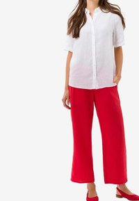 Femme portant une chemise blanche à boutons et à manches courtes, un pantalon rouge à jambes larges et des chaussures à talons bas rouges, se tenant avec une main dans la poche.