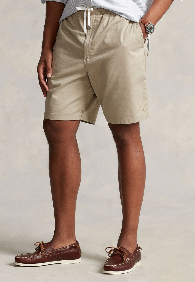 Chaki spalvos medvilniniai šortai su elastinga juosmens juosta, dekoratyviniu baltu raišteliu ir šoninėmis kišenėmis. Derinami su rudos spalvos laivo tipo batais su raišteliais.