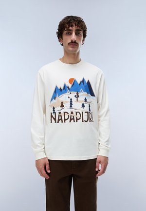 Un homme porte un sweat-shirt Napapijri couleur crème orné d’un motif coloré représentant une montagne et une forêt, associé à un pantalon marron foncé.