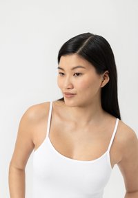 Jeune femme aux longs cheveux noirs portant un haut sans manches blanc, regardant légèrement vers la gauche devant un fond clair et uni.