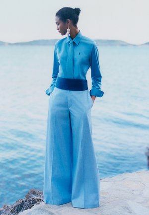 Naine laia säärega helesinistes pükstes ja sobiva lipsuga särgis seisab kivilise ranna ääres ning vaatab vasakule üle rahuliku sinise vee.