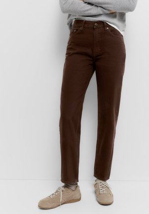 Person iført brune bukser med høj talje, grå sweater, beige sneakers, står med armene korsede mod en hvid væg.