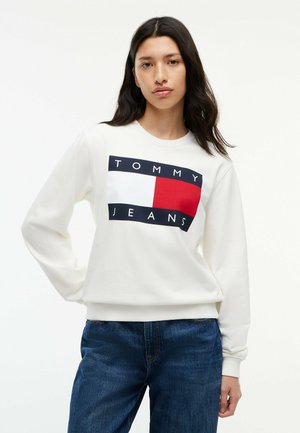 Młoda kobieta o długich ciemnych włosach, ubrana w białą bluzę Tommy Jeans i niebieskie jeansy, stojąca z jedną ręką na biodrze.