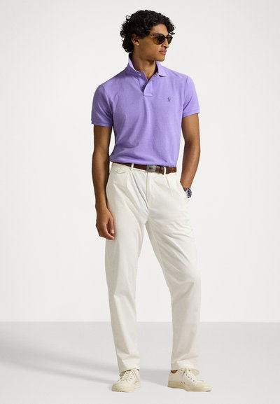 Πολο πουκάμισο από βαμβάκι σε λεβάντα, κοντά μανίκια, με κουμπιά δύο θέσεων, συνδυασμένο με λευκά παντελόνια, καφέ ζώνη και ανοιχτόχρωμα αθλητικά παπούτσια.