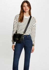 Schwarze Leder-Crossbody-Tasche mit runder Form, goldenen Beschlägen, Kontrastnähten und einer Klappenverschluss. Gestreifter Strukturpullover und blaue Jeans.