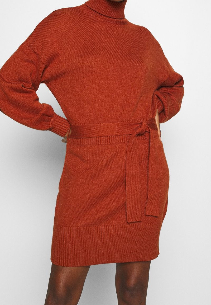 Femme portant une robe-pull en maille couleur rouille, manches longues, ceinture nouée à la taille et ourlet côtelé, debout, les mains sur les hanches.