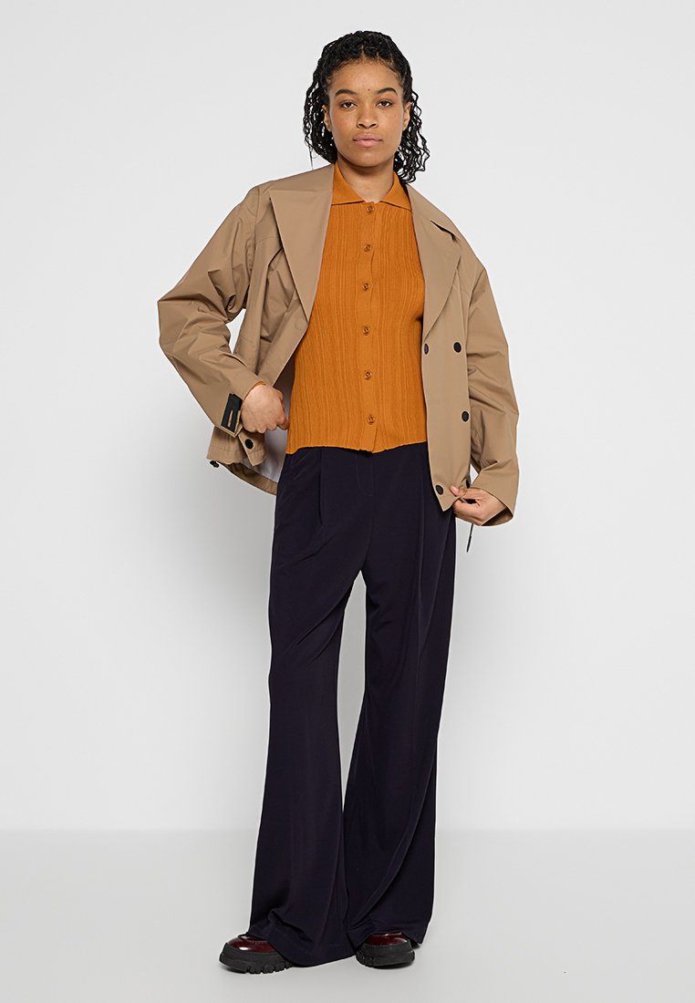 Tan wasserfeste Jacke mit doppelreihiger Knopfleiste über einem orangefarbenen, gerippten Hemd. Marineblaue weite Hose; dunkle Schuhe.