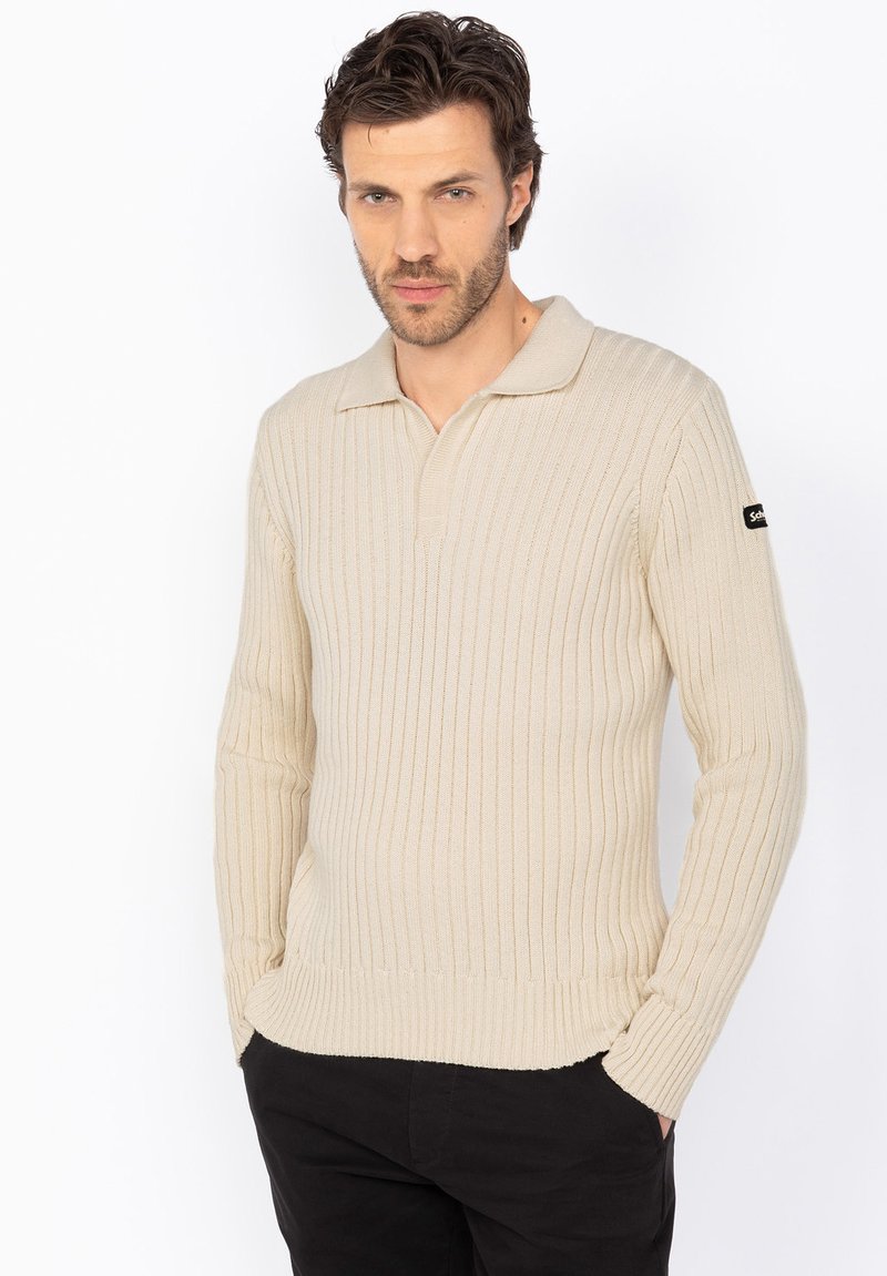 Beige ribstrikket pullover med lange ærmer, polo-krave og et logo-patch på venstre ærme. Glat tekstur og tætsiddende design.