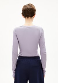 Person mit kurzen braunen Haaren, die sich abwendet, und ein helllila langärmliges Shirt sowie eine dunkelblaue Hose mit Elastikbund trägt.