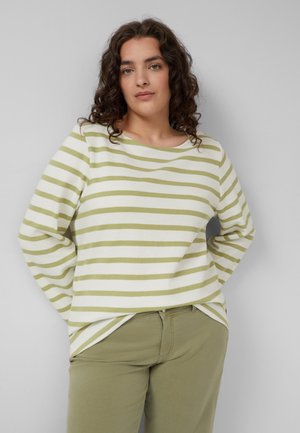 Kobieta z kręconymi włosami, ubrana w zielono-białą, w paski, bluzkę z długim rękawem oraz oliwkowo-zielone spodnie, stojąca na jednolitym jasnoszarym tle.