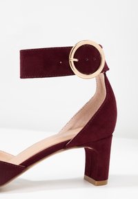 Burgundy mockal lädersandalett med ankelrem och klack, försett med ett guldspänne och en blockklack. Har en slät innersula och en öppen tådesign.