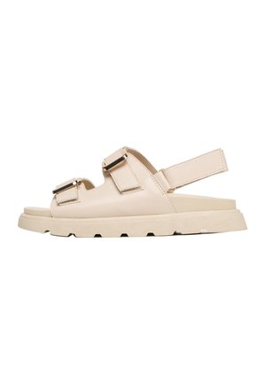 Beige åben tå sandal med to justerbare spænder over foden og en ankelrem med velcrolukning, flad sål med mønster.