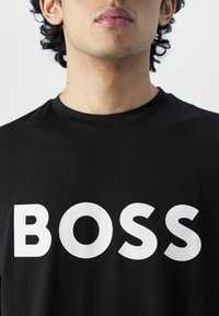 Melnā kokvilnas T-krekls ar izteiktu baltu tekstu, kas izsaka "BOSS". Arapa kakla dizains, gluda tekstūra un brīvs piegriezums ar minimāliem šuvju detalizējumiem.