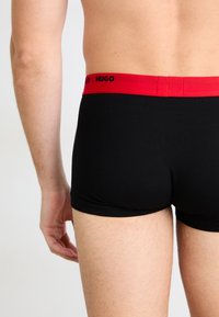 Schwarze Baumwoll-Boxershorts mit einem roten Elastikbund, auf dem der Markenname "HUGO" in Schwarz steht. Glatte Textur und enganliegendes Design.