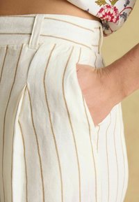 Pantaloni de culoare crem cu dungi verticale maro, cu față plată și buzunare laterale, confecționați dintr-un material texturat pentru o croială lejeră.