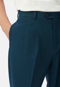 Op maat gemaakte broeken van donker teal stof; heeft een vlakke voorkant, zijzakken en een enkele knoopsluiting bij de taille. Glad van textuur.