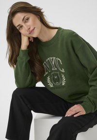 Donna con capelli lunghi e castani che indossa una felpa verde di New York e pantaloni neri, seduta con la testa appoggiata sulla mano su uno sfondo bianco.