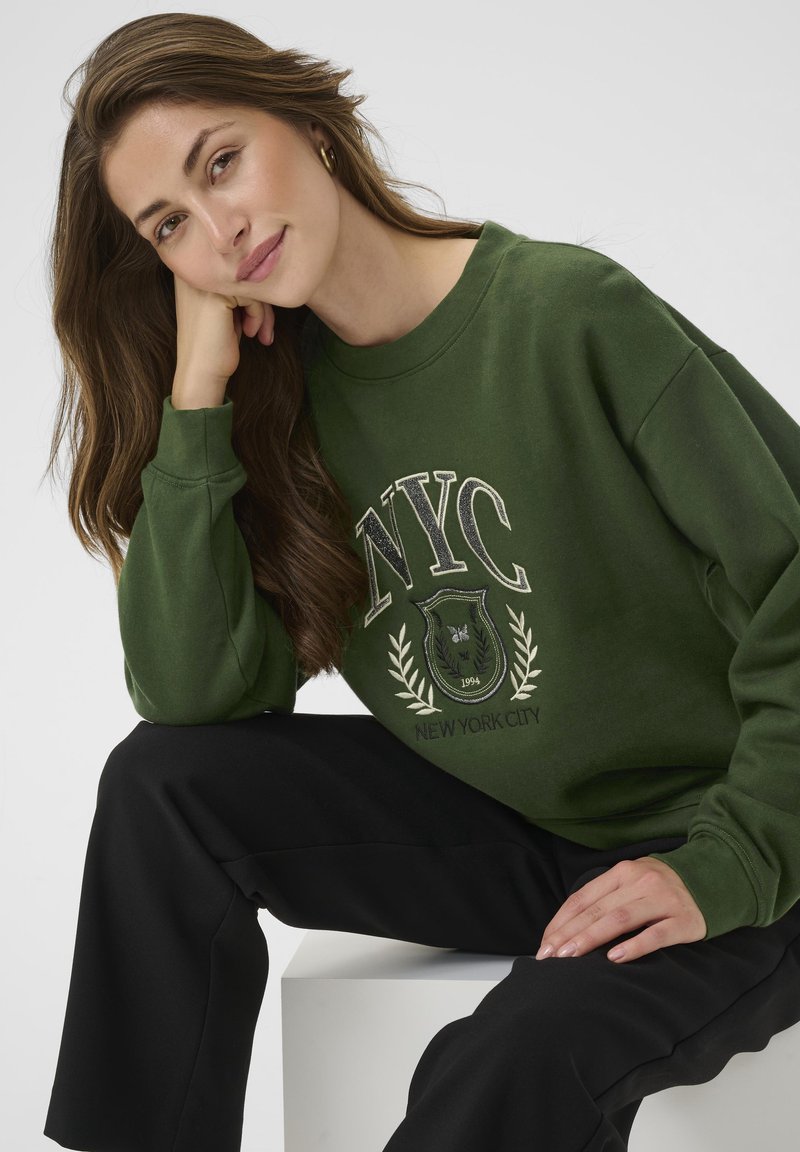 Donna con capelli lunghi e castani che indossa una felpa verde di New York e pantaloni neri, seduta con la testa appoggiata sulla mano su uno sfondo bianco.