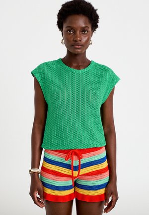 Donkere vrouw die een groene gebreide mouwloze top draagt en veelkleurige gestreepte gebreide shorts met een rode trekkoord.