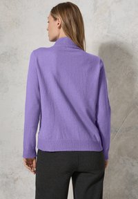 Pull en tricot violet avec un col montant et une finition côtelée aux bords et aux poignets. Texture douce, coupe décontractée. Porté avec un pantalon gris foncé.