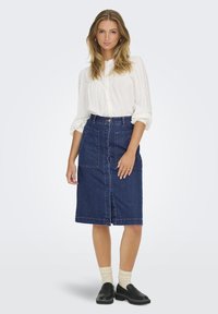 Lahka, bela bluza s pikčastim vzorcem, vtaknjena v jeans pencil krilo z dvema sprednjima žepoma in gumbom na sredini spredaj.