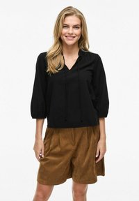 Blusa nera, dal taglio morbido, con scollatura a V, dettaglio con cordoncino e maniche a sbuffo tre quarti, abbinata a pantaloni corti marroni, morbidi al tatto.
