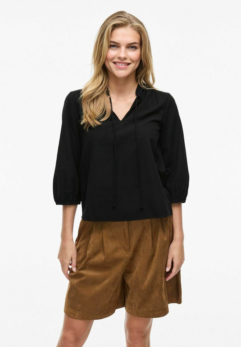Blusa nera, dal taglio morbido, con scollatura a V, dettaglio con cordoncino e maniche a sbuffo tre quarti, abbinata a pantaloni corti marroni, morbidi al tatto.
